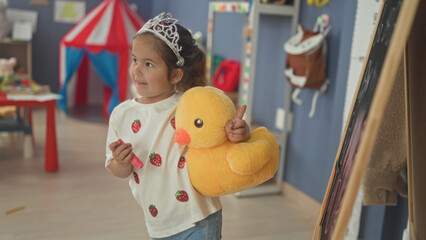 Young girl wearing tiara holds pink marker and large yellow plush duck, points marker to easel near toy table in an indoor playroom building  playful joy. © Krakenimages.com