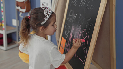 Girl drawing on chalkboard, hand holding pink chalk while sketching letters in preschool classroom  curiosity learning imagination. © Krakenimages.com