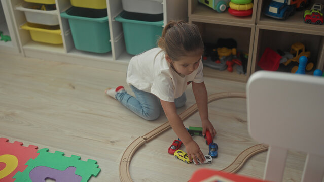 Hispanic girl kneels and reaches arranging toy cars on a wooden train track beside foam alphabet mat and storage shelves inside a playroom building; concentration imagination play.