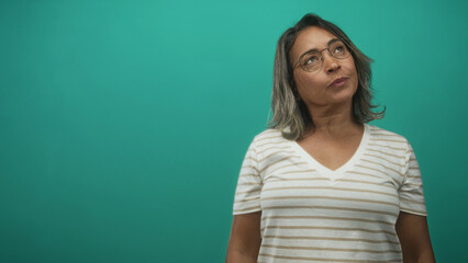 Fototapeta premium Middle aged hispanic woman in a striped v neck shirt wearing glasses, head tilted and eyes closed against a teal studio backdrop; quiet contemplation.