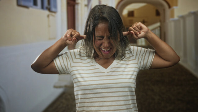 Woman in striped shirt plugs ears with fingers and grimaces inside a building corridor with archway and balustrade; irritation discomfort.
