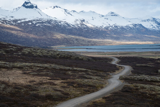 Winding gravel road crossing a rugged landscape with snow capped mountains and a fjord. Iceland Icelandic landscape