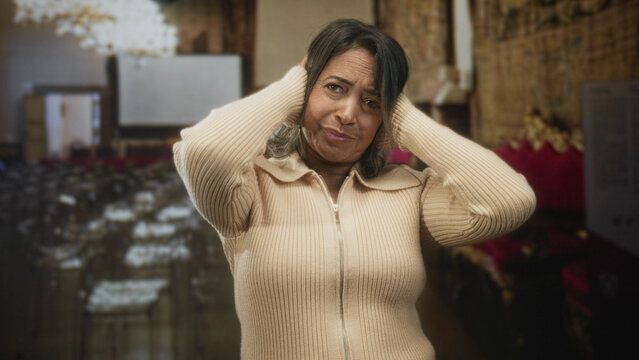 Woman presses hands to ears and winces, standing among rows of chairs and gilded seating in an ornate hall building; distress sensory overload.