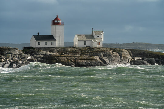 Reliability at the maritime frontier is expressed by the clear white form of Langoytangen lighthouse above restless green water and rugged rock in Bamble, Norway.