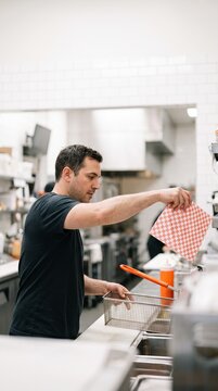 A male cook in a commercial restaurant kitchen prepares food using a fryer basket and checkered paper