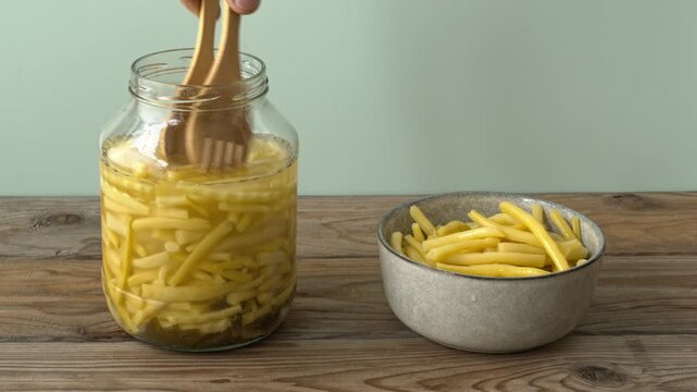 Picking fermented yellow wax beans from a jar with wooden tongs. Organic homemade sour Golden Teepee bean pickles are being transferred to a ceramic bowl. Copy space.