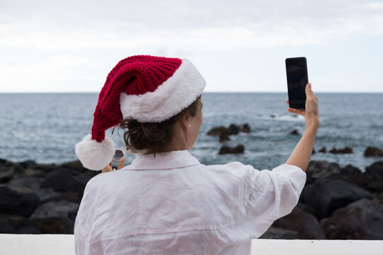 Rear view of a woman wearing a red Santa hat and white shirt taking a selfie with her smartphone near a rocky sea coast.