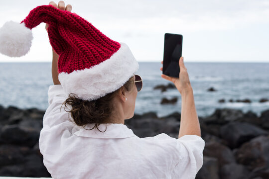 Rear view of a female holding her Santa hat and taking a mobile photo in front of a rocky coastline and sea.