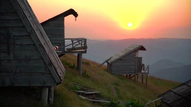 Glowing Sunset Above Rustic Cabins in Carpathian Mountains
 Warm evening sun glowing above distant mountain ridges and traditional wooden shepherd cabins on a quiet Carpathian hillside.