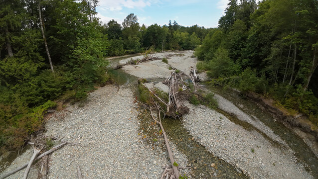 An aerial wide shot looks along a shallow rocky river bed filled with fallen driftwood logs, surrounded by dense green forest trees under a bright blue sky with clouds.