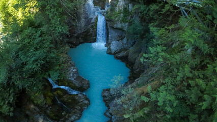 A high angle wide shot looks straight down over Mamquam Falls plunging into a bright milky turquoise glacier water pool inside a deep rocky canyon surrounded by dense forest trees. © Matt