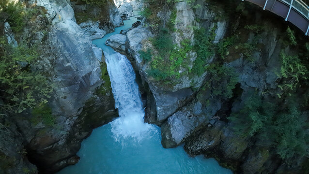 A high angle wide shot looks directly down into a deep rocky cliff canyon where Mamquam Falls plunges heavily into a rushing stream of milky turquoise glacier river water.