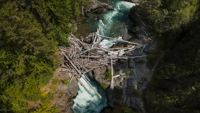 A top down close up aerial shot captures an extensive log jam of weathered driftwood logs blocked between large canyon boulders across the rushing turquoise Cheakamus River.