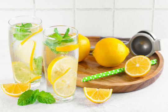 Refreshing homemade lemonade in glasses with fresh lemon slices, mint leaves, ice cubes, sparkling water served on wooden tray on white tiled kitchen background. Summer citrus beverage, drink concept.