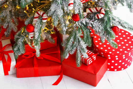 Beautiful presents, gift boxes wrapped in elegant red paper under green snowy festive Christmas tree with warm lights. Cozy winter holiday atmosphere perfect for seasonal celebrations and decor themes