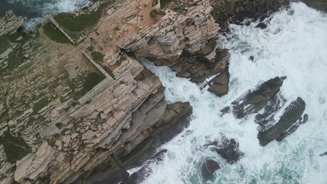 Aerial view of Baleal Island featuring rugged limestone cliffs and powerful white Atlantic waves crashing against the rocky coastline in Peniche, Leiria District, Portugal.