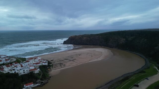 Aerial view of Odeceixe beach where the river meets the Atlantic Ocean, featuring white buildings on the cliffs under a cloudy sky in Odeceixe, Faro District, Portugal.