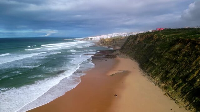 Aerial view of the sandy Praia de Sao Juliao beach with dramatic cliffs, crashing waves, and a vast ocean under a cloudy sky in Sao Joao das Lampas, Lisboa, Portugal.