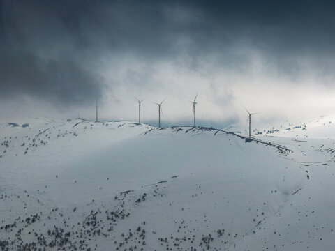 Aerial view of wind turbines standing on a snow-covered mountain ridge under dramatic dark clouds in Anversa degli Abruzzi, Abruzzo, Italy.