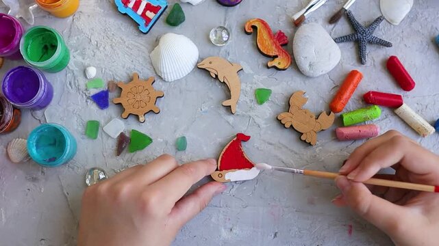 Top view of children's hands painting small wooden sailboat with brush, surrounded by art supplies, sea shells, stones, and nautical shapes on textured gray background