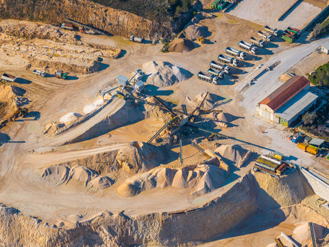 Aerial view of a sand and gravel quarry featuring conveyor belts, heavy machinery, and a line of parked trucks in Capestrano, Abruzzo, Italy.