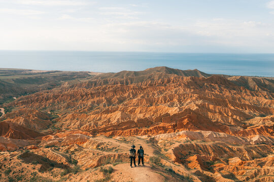Tosor, Kyrgyzstan - 16 June 2026: View of two hikers standing on a ridge overlooking the red rock formations of Skazka Canyon with the blue waters of Issyk-Kul Lake in the distance.