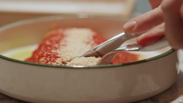 Close up of hand using fork and knife to cut into food covered in red sauce with grated cheese revealing texture details. scene is set on kitchen countertop during preparation process