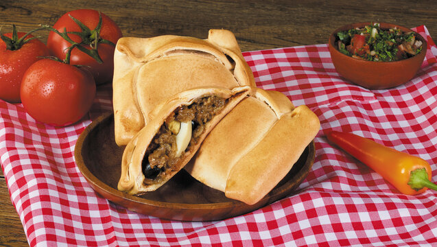 Baked Chilean beef empanadas filled with minced meat, hard-boiled egg, and olives, served on a wooden plate with pebre sauce, fresh tomatoes, and chili pepper over a red checkered tablecloth