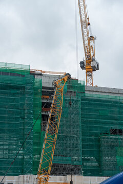 Upward perspective of a massive high-rise building project shrouded in green protective scaffolding nets, operated by two large industrial cranes against a cloudy sky.
