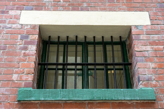 Symmetrical exterior view of vertical black security bars over a green framed window set in a weathered red brick institutional building wall.