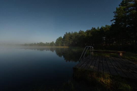 Tranquil wooden swimming pier with ladder extending into a calm misty lake in Omma bog, Estonia, under a dark starry night sky