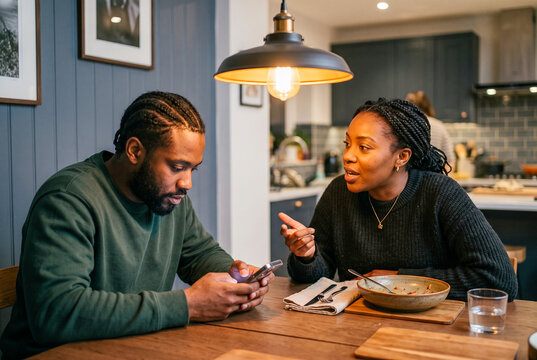 Young Black woman trying to talk to her distracted partner who is looking at his phone during dinner at home