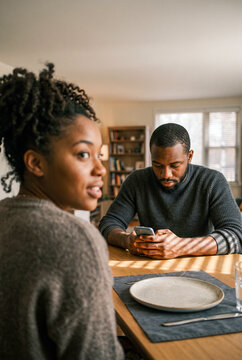 Young Black couple sitting at dining table while man is distracted by his smartphone and woman looks concerned