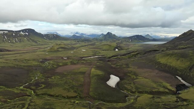 Aerial drone view with a push in and pan left reveals the expansive Laugavegur hiking trail valley on Iceland. Lush green mountains, a winding path, distant lake, and snow patches under a cloudy sky.