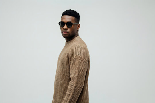 Portrait of a young black man wearing black sunglasses and a brown ribbed knit sweater standing against a plain light gray studio background
