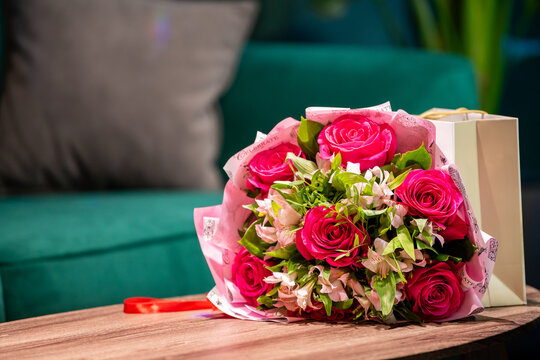 Bouquet of pink roses and white flowers with gift bag on wooden table at home. Floral present for celebration. Valentine day, mothers day, birthday or womens day gift concept