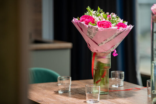 Bouquet of pink roses in glass vase on wooden table with empty glasses. Flower gift for holiday celebration, birthday or Valentine day. Home interior decoration concept.