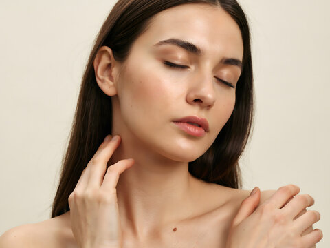 Portrait of a young brunette woman with closed eyes, touching her neck and shoulder, showcasing glowing skin against a neutral light background