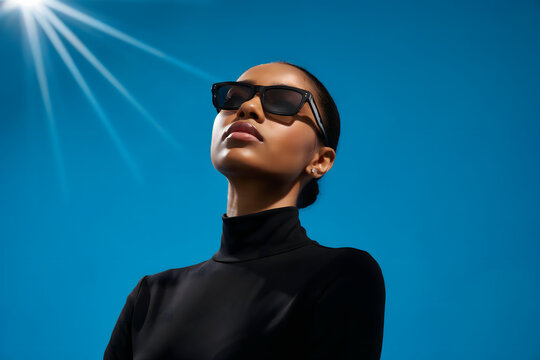 Young woman wearing black sunglasses and a black turtleneck against a bright blue sky with a visible sun flare in the upper left corner