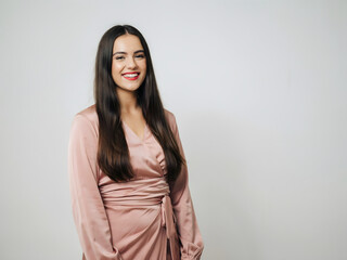 Obraz premium A smiling young woman with long dark hair wearing a dusty pink satin wrap dress standing against a plain light gray studio background