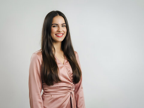 A smiling young woman with long dark hair wearing a dusty pink satin wrap dress standing against a plain light gray studio background