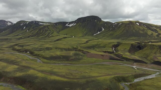 Aerial drone shot panning left over the Laugavegur hiking trail on Iceland, revealing stunning rugged green mountains, a winding braided river, and a vast, remote volcanic landscape.