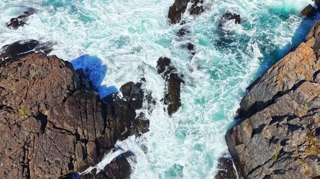 Waves roll in and out, hitting the rocks along the shore. The scene shows deep blue water and foam surrounding the rugged cliffs. Light from the sun reflects on the surface of the ocean.