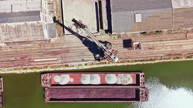 A barge is moored at a dock while a crane lifts sand from its deck. The surrounding area has buildings and tracks in daylight. Water reflects the scene.