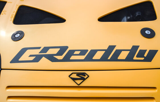 Close-up of yellow car hood featuring black GReddy decal two mesh covered vents secured with quick release hood pins and stylized black "S" emblem in diamond shield below