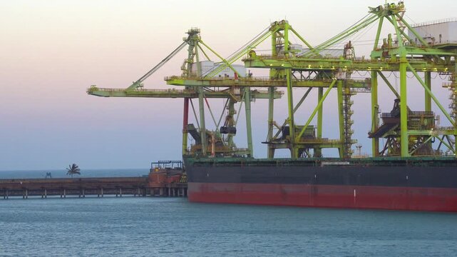 Side view of a large bulk carrier vessel berthed at a commercial sea port terminal, actively unloading cargo with giant green ship unloader cranes against a calm sunset sky