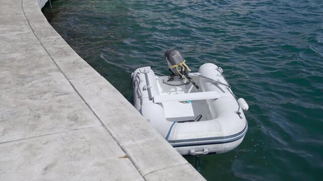 A white inflatable dinghy fitted with a small outboard motor floats quietly on calm sea water next to a concrete pier, moored and empty.
