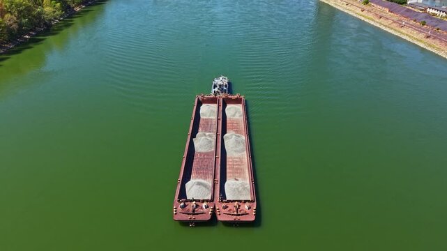 Two large barges move on a river, carrying loads of sand. The scene shows clear water and banks lined with trees. Sunlight reflects on the water surface.