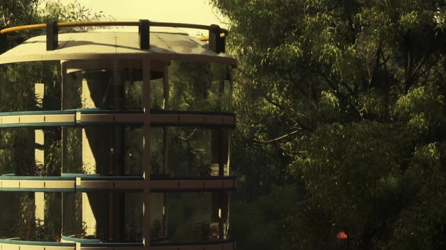 A glass structure is located in an area surrounded by trees. It appears to be well designed, blending with the environment. The sun is shining on the building, highlighting its features.