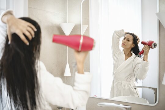 A woman is using a hair dryer to style her wet hair while standing in front of a large mirror. The bright bathroom features minimalistic design elements and soft natural light filtering through